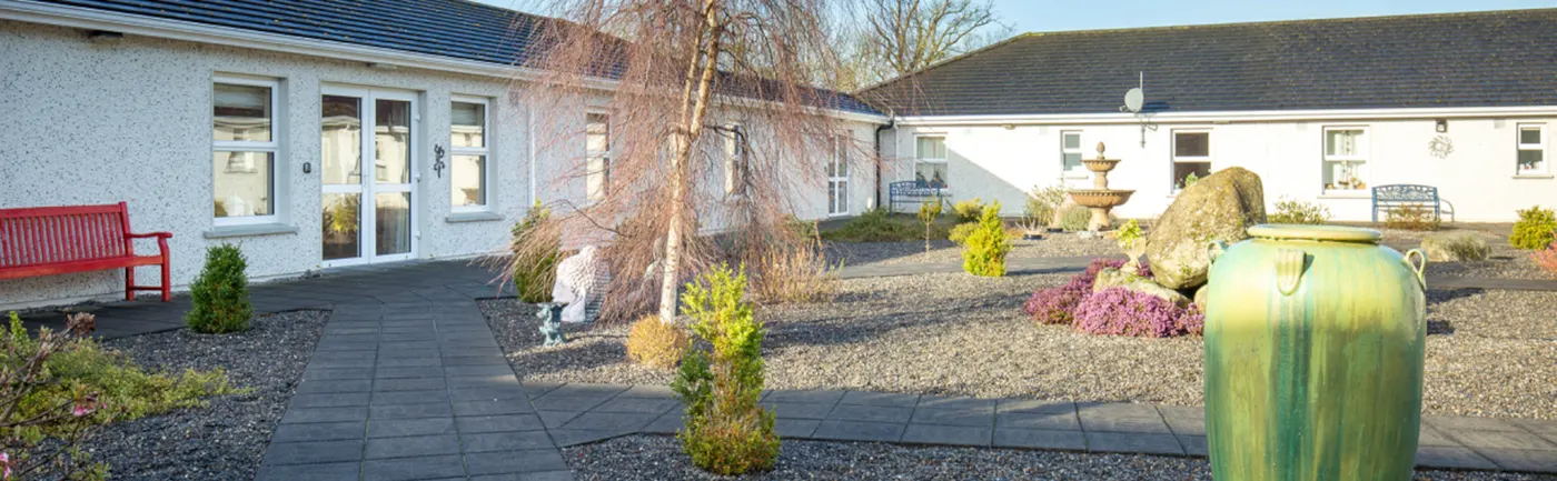 Beechwood Nursing Home, Leighlinbridge, Co. Carlow: single-storey pebble-dashed building around a gravel courtyard garden with a red bench, birch tree, fountain and large green urn, no people; page banner on the Beechwood accommodation page, which describes a ground-floor home with three secure gardens