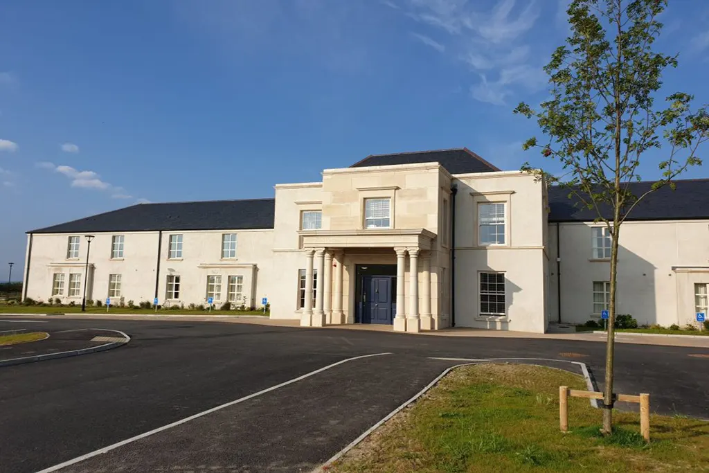 Castlebridge Manor Nursing Home, Castlebridge, Co. Wexford: front of the two-storey cream building with its columned entrance porch, car park and young tree, blue sky, no people; site alt text: Exterior shot of Castlebridge Nursing Home, Co. Wexford