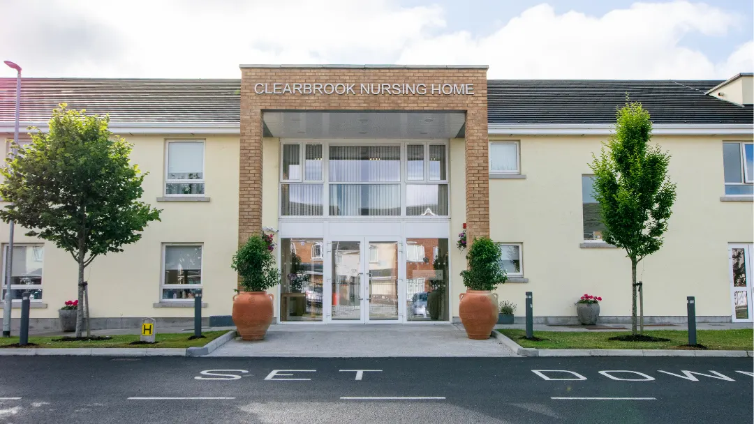 Clearbrook Nursing Home, Finglas West, Co. Dublin: front entrance with the Clearbrook Nursing Home sign over the glazed porch, two trees and planters, set-down bay in front, no people; site alt text: Exterior of front of Clearbrook Nursing Home, Finglas, Dubin 11