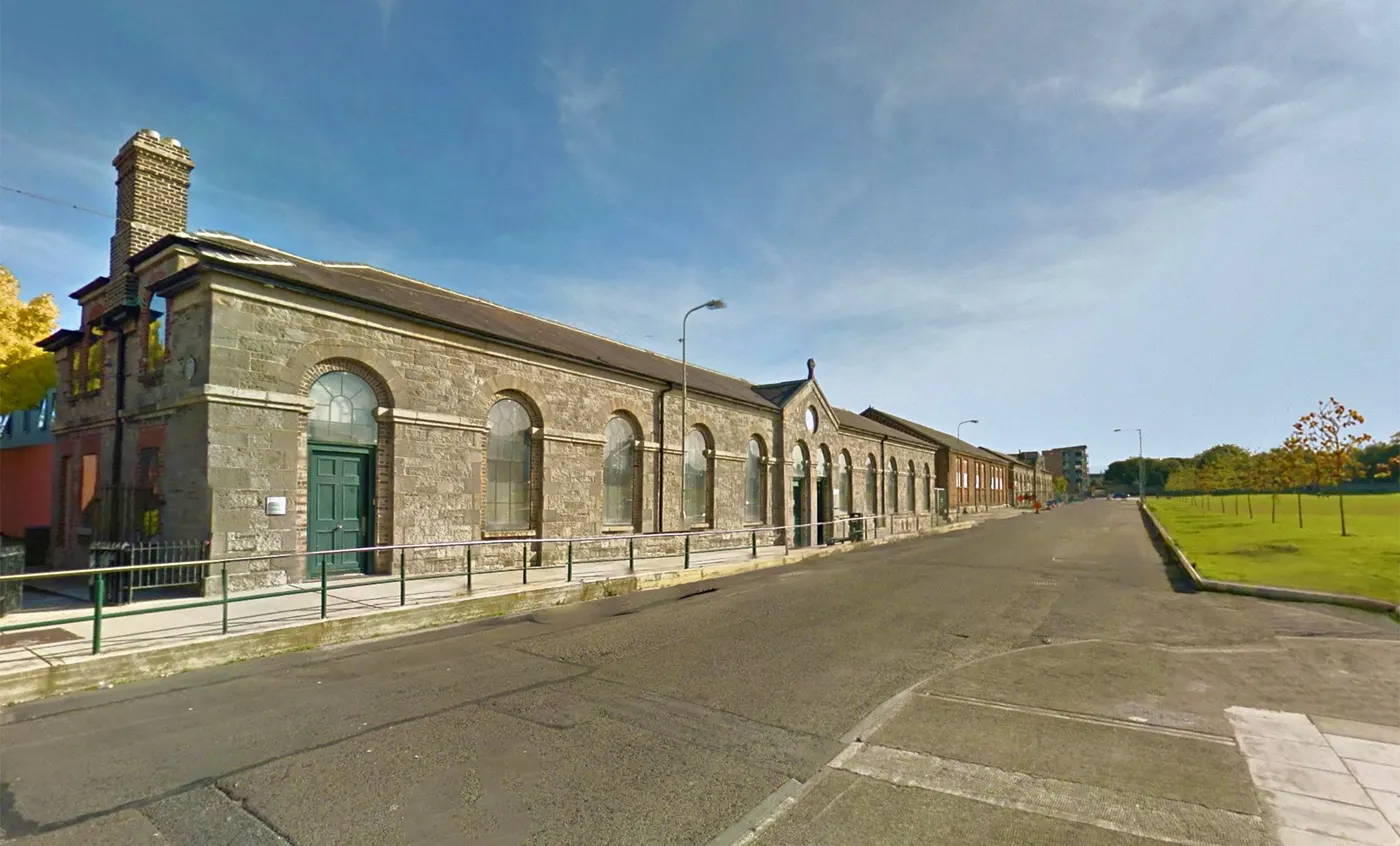 Inchicore Family Doctors, Inchicore, Co. Dublin: Street-level view of a long single-storey cut-stone building with arched windows and a green door beside a road and green.